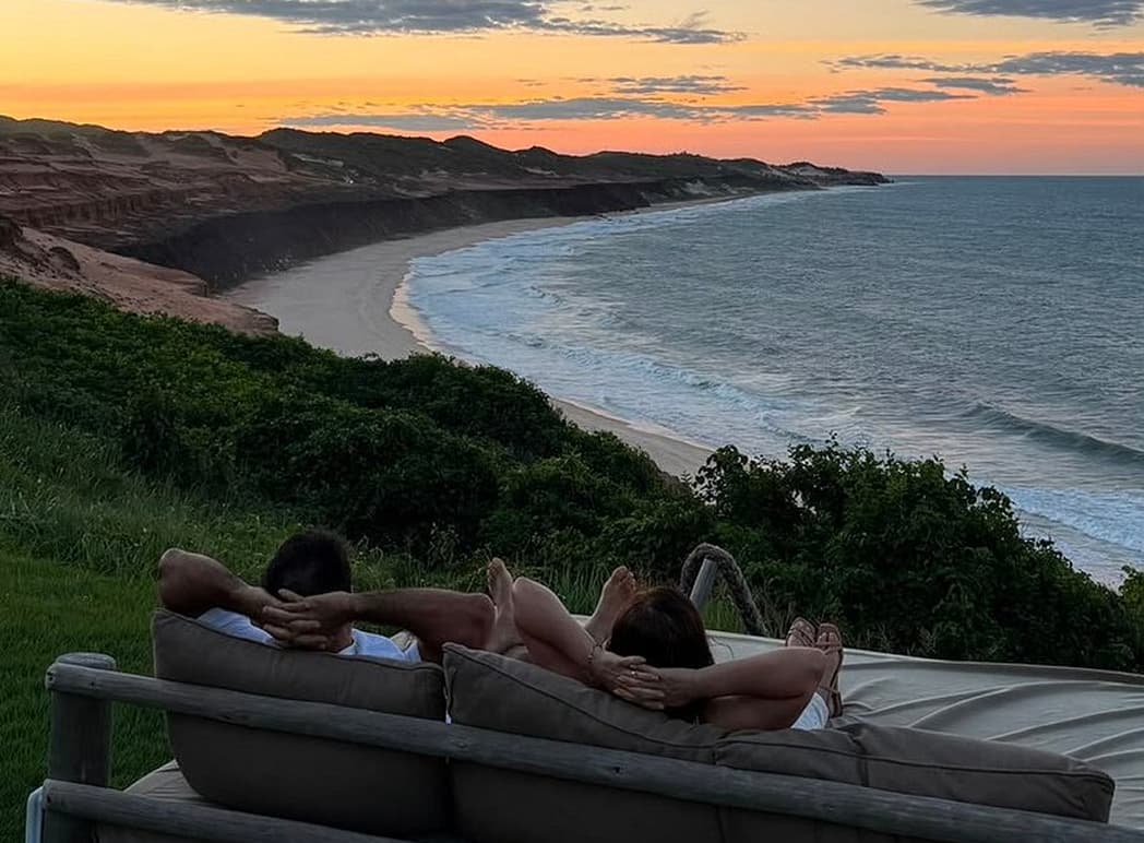 Casal deitado em cama externa observando o pôr do sol sobre as falésias e o mar de Sibaúma