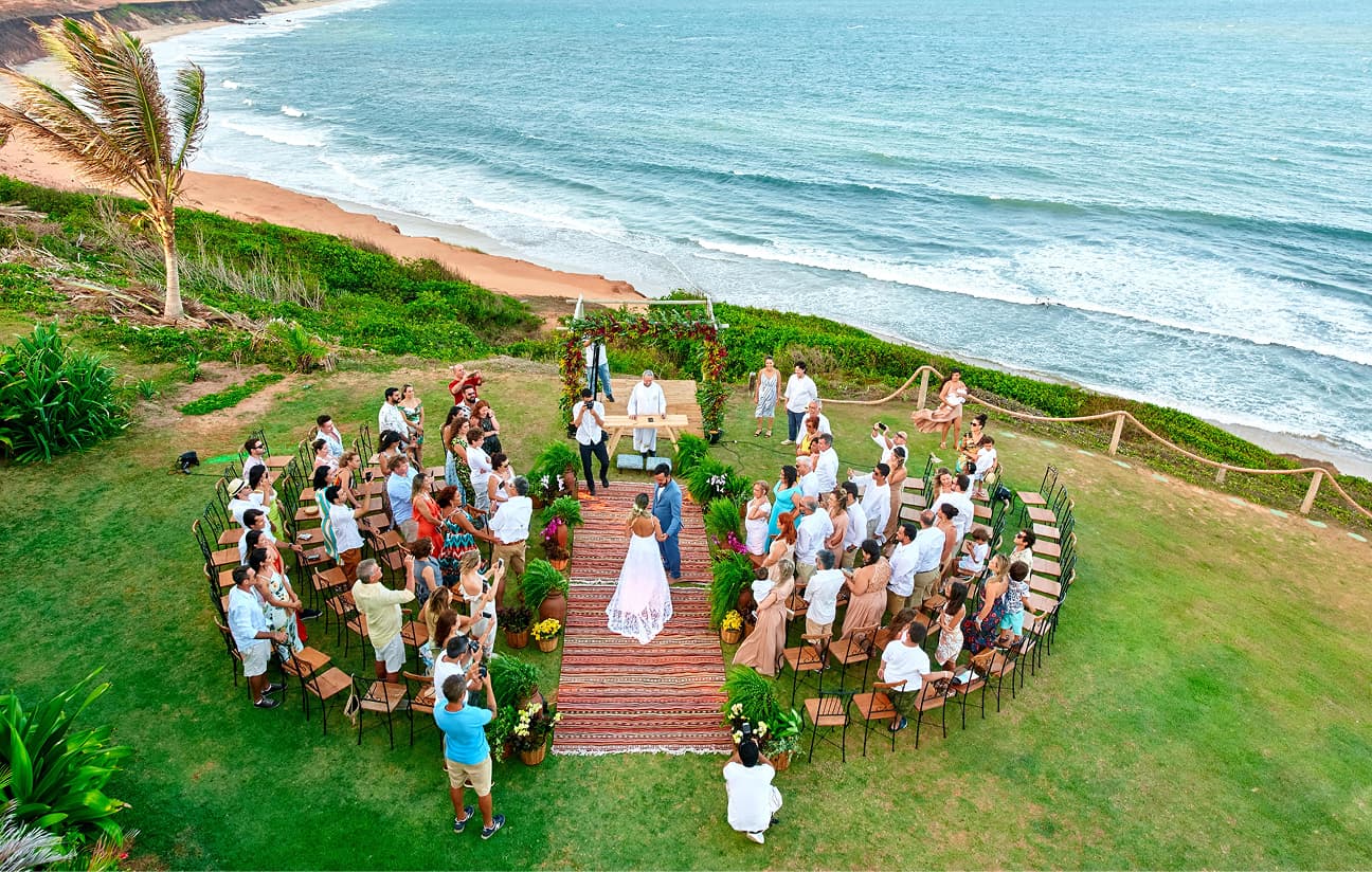 Vista aérea de um casamento ao ar livre em um gramado à beira-mar, com convidados dispostos em semicírculo ao redor dos noivos.