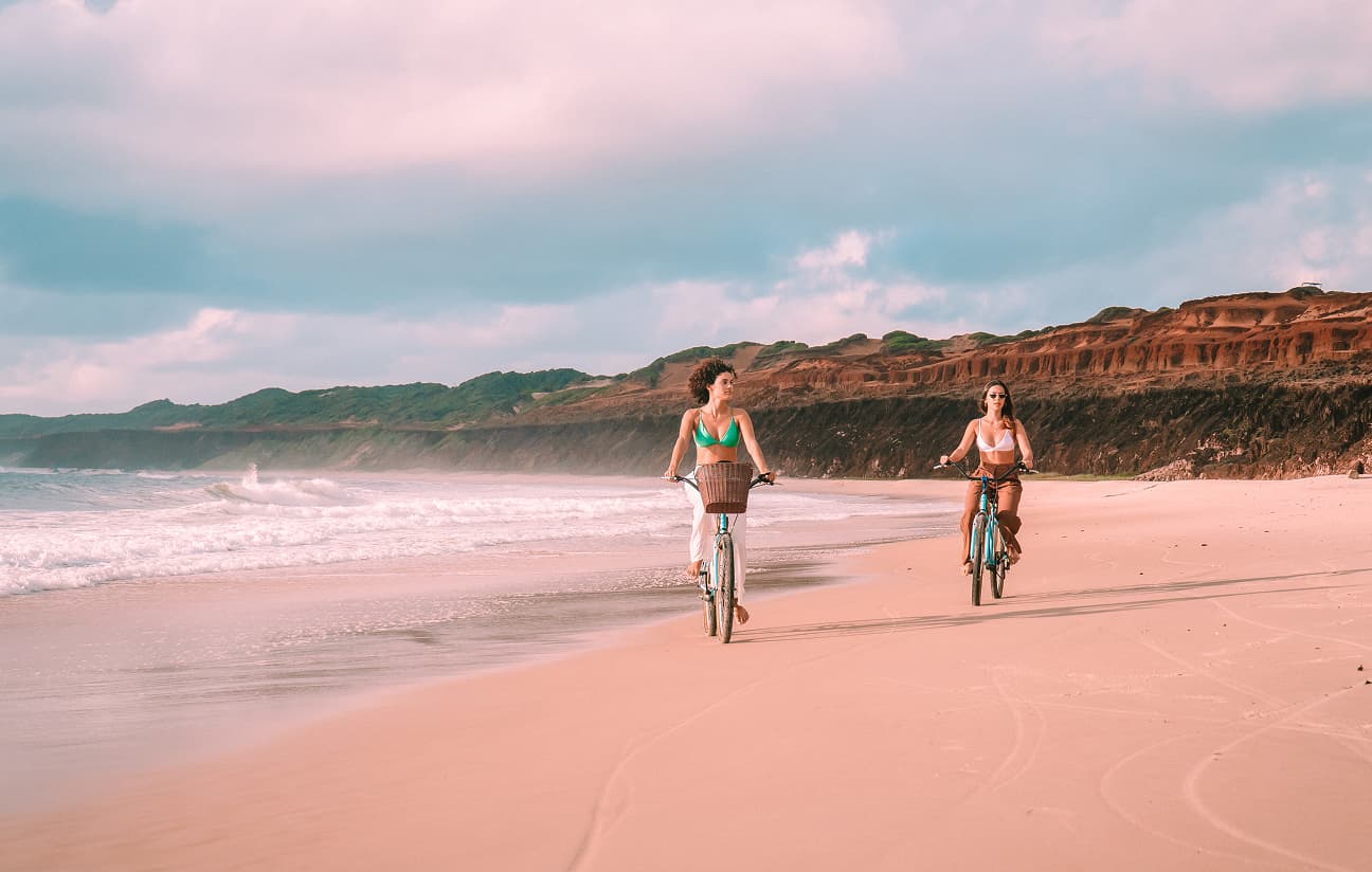 Duas mulheres andam de bicicleta em uma praia de areia clara, com falésias avermelhadas ao fundo sob um céu nublado.