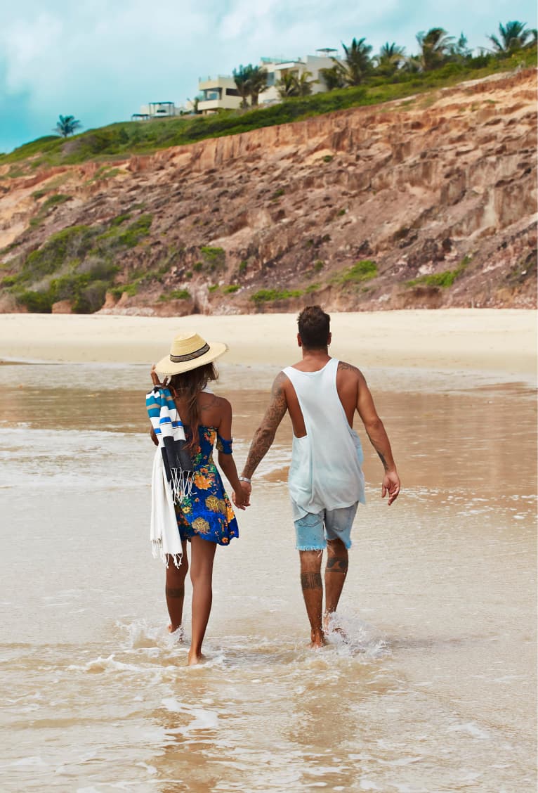 Casal caminhando de mãos dadas na areia da praia de Sibaúma com as falésias e o Kilombo Villas ao fundo