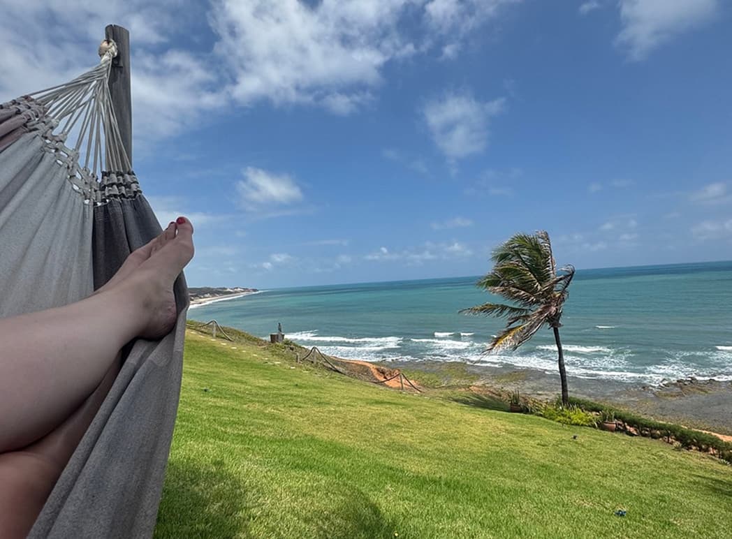 Pés apoiados em rede com vista para o mar turquesa e coqueiro na praia de Sibaúma