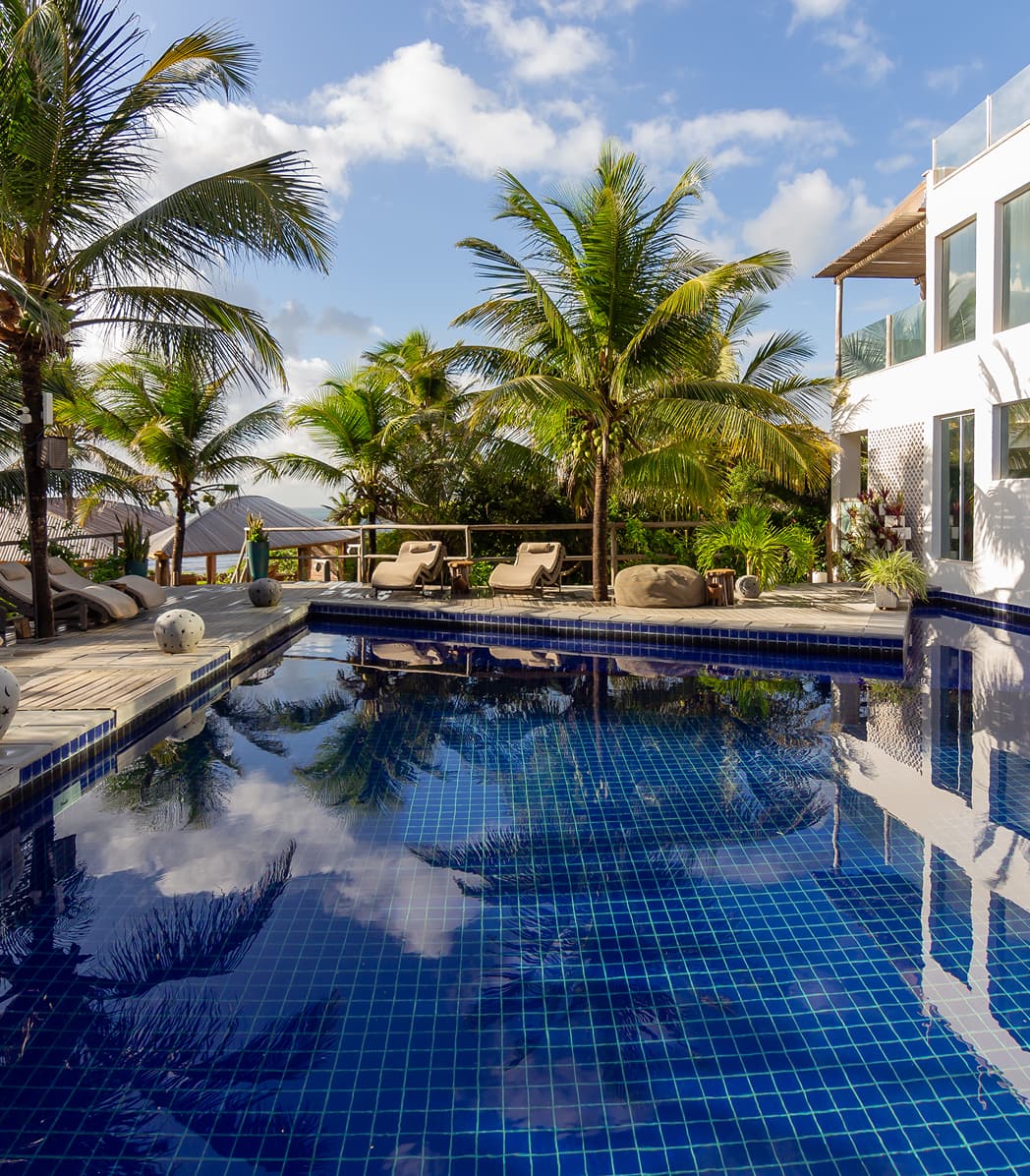 Piscina azulejada com coqueiros e villa refletidos na água em dia de céu azul no Kilombo Villas