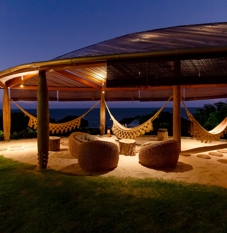 Pavilhão de palha com redes e pufes de fibra natural iluminados ao anoitecer, de frente para o mar, no Kilombo Villas