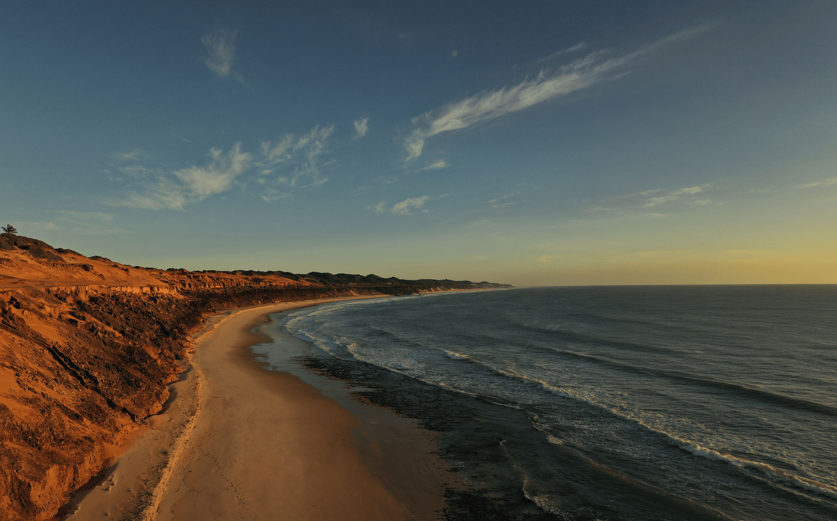Vista aérea da costa de Sibaúma com falésias avermelhadas, praia dourada e oceano ao pôr do sol