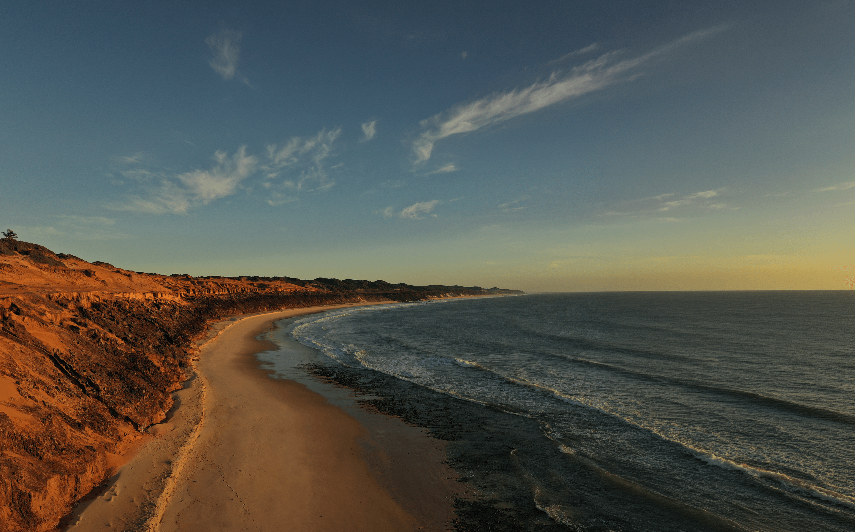Vista aérea da costa de Sibaúma com falésias avermelhadas, praia dourada e oceano ao pôr do sol