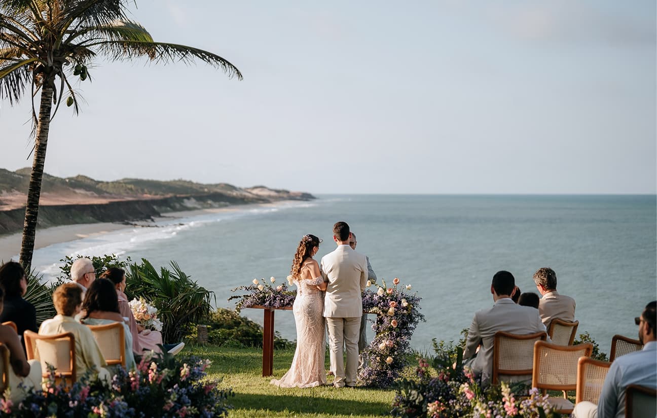 Cerimônia de casamento ao ar livre em um gramado à beira-mar, com os noivos de frente para o oceano e convidados sentados em cadeiras de palha.
