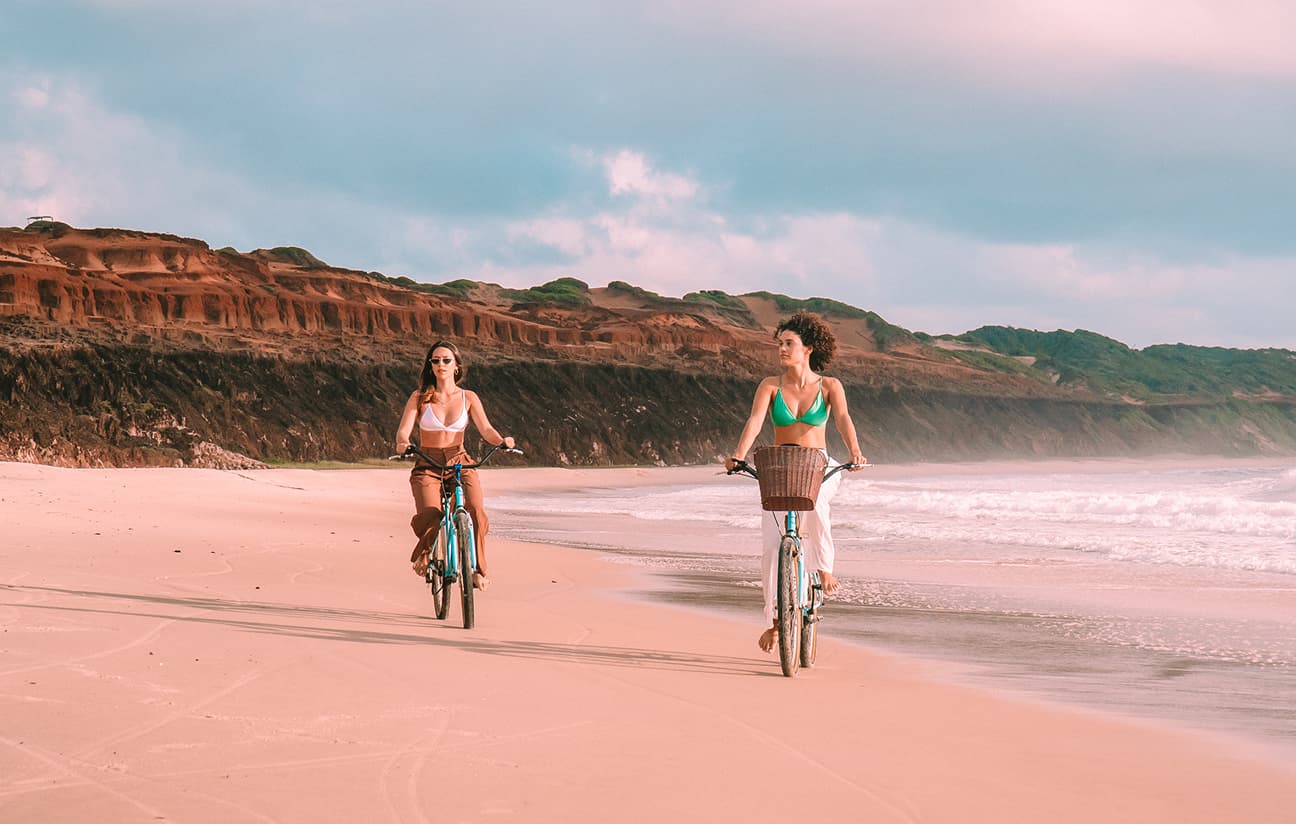 Duas mulheres andam de bicicleta em uma praia de areia clara, com falésias avermelhadas ao fundo sob um céu nublado.