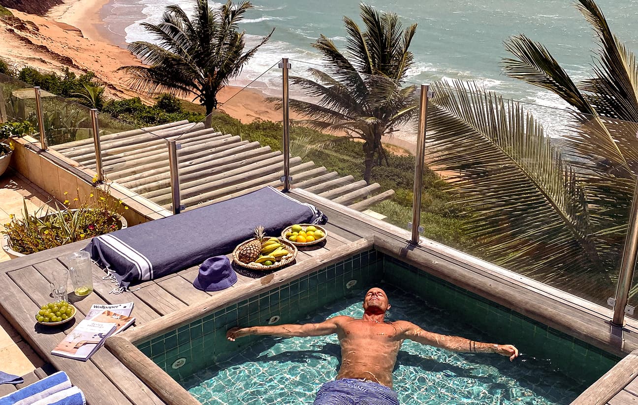 Homem relaxa boiando em uma pequena piscina privativa de um deck elevado, com vista panorâmica para o mar e coqueiros.