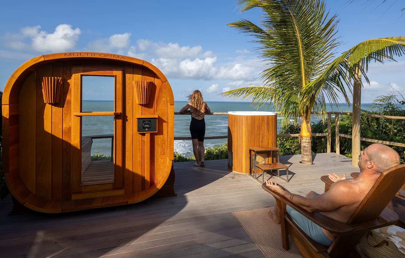 Sauna de madeira em um deck com vista para o mar, onde uma mulher admira o horizonte e um homem relaxa em uma espreguiçadeira sob coqueiros.
