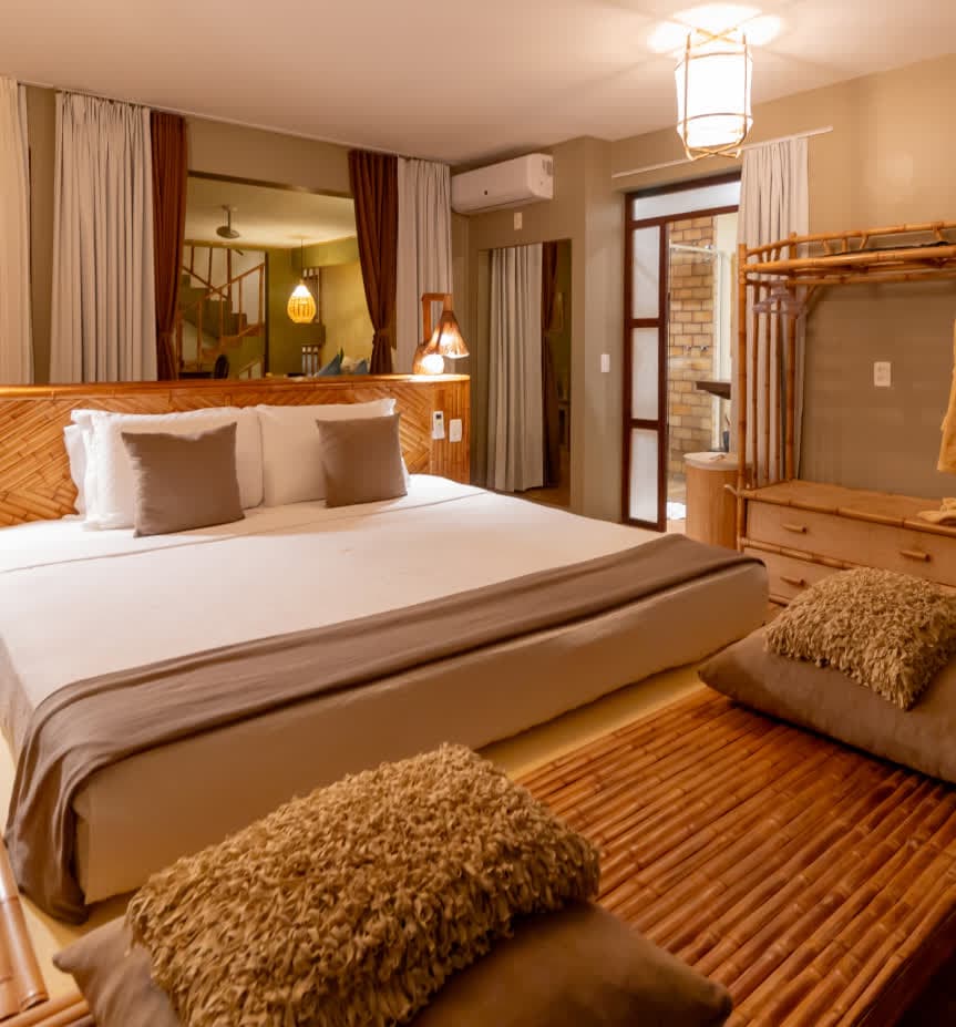 Kilombo Villas - Sibraáma Ocean - Quarto principal da suíte com uma ampla cama king size decorada com cabeceira de bambu. À frente da cama, há um banco de bambu com almofadas felpudas em tons terrosos. O ambiente possui iluminação suave e móveis de materiais naturais.