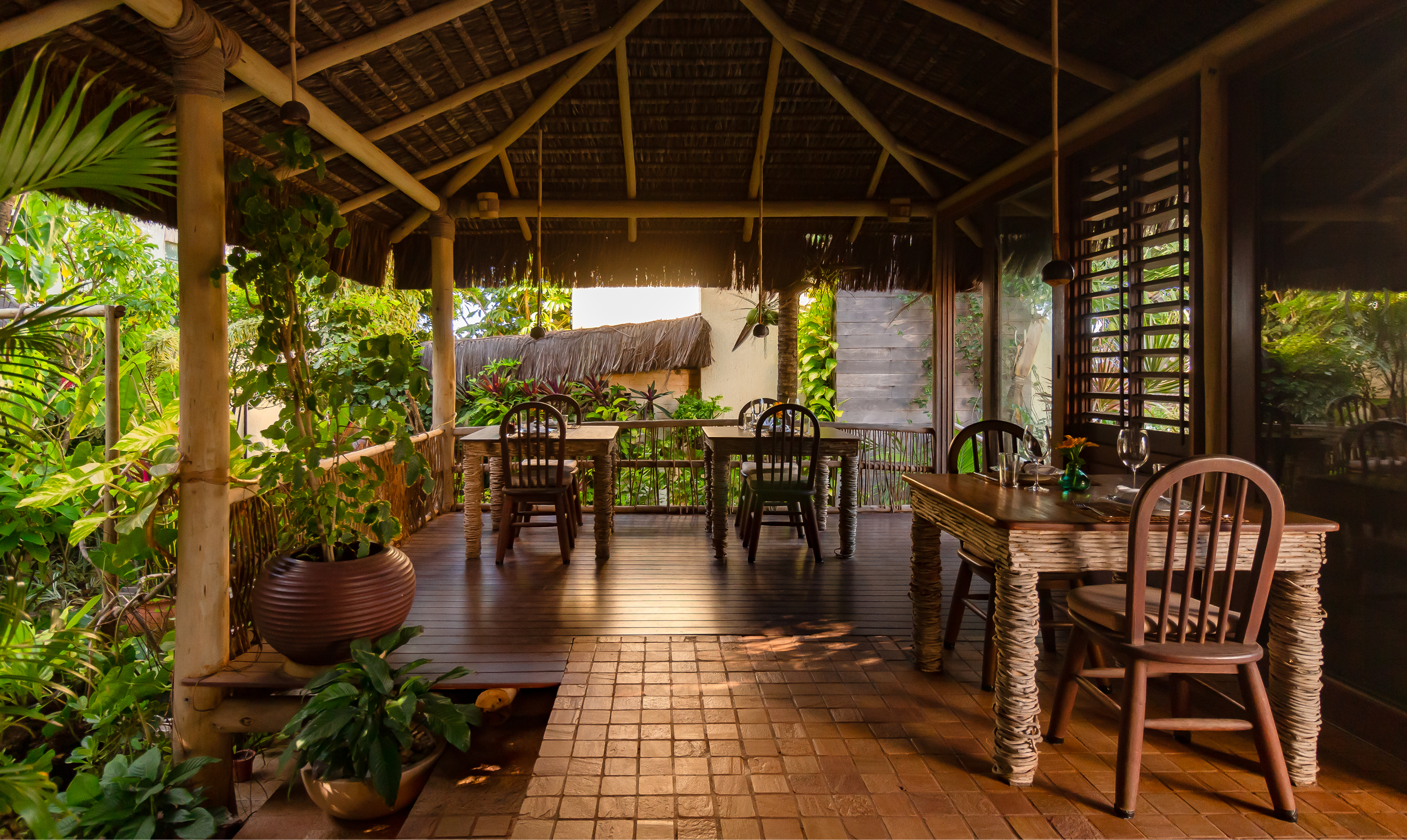 Salão aberto do Kibo Restaurante com mesas de madeira, plantas tropicais e teto de palha no Kilombo Villas