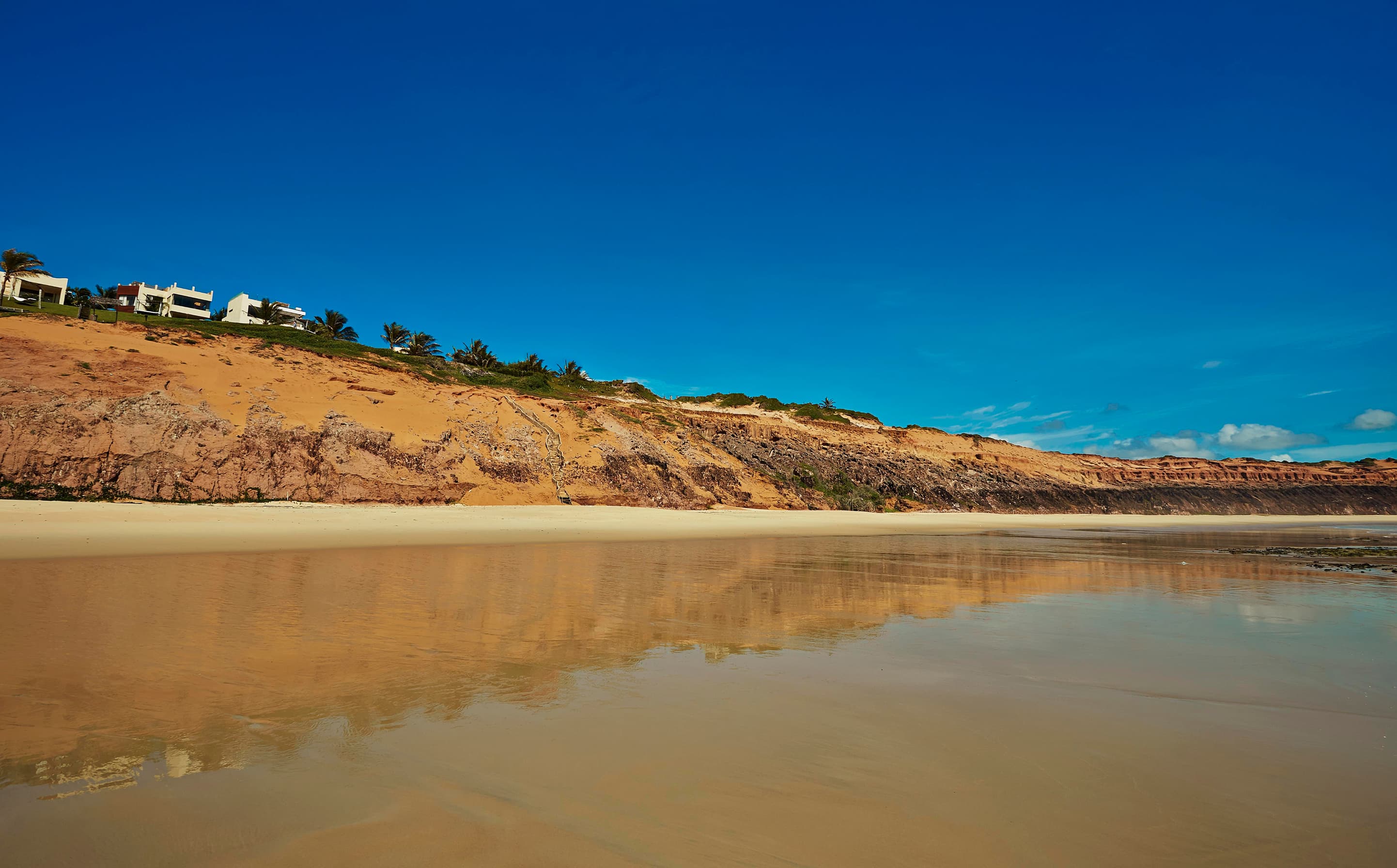 Praia de Sibaúma com falésias avermelhadas, areia molhada e reflexo do céu azul, Tibaú do Sul