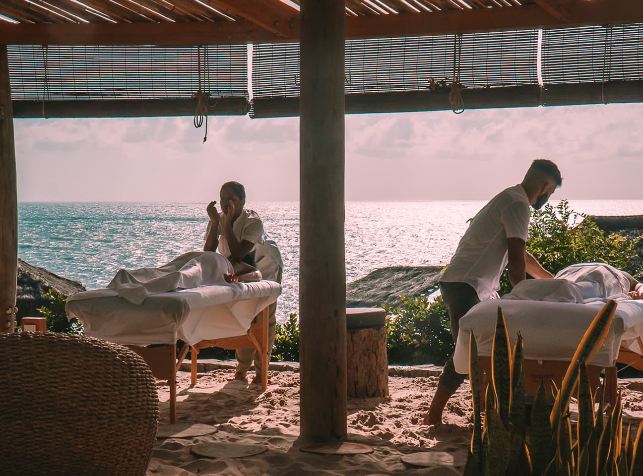 Duas massagens simultâneas em macas sobre a areia, sob um pergolado de bambu, com o mar ao fundo