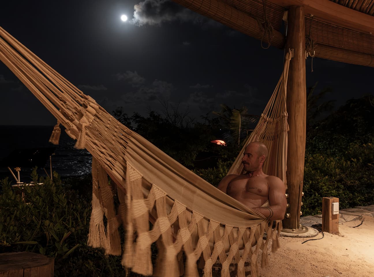 Hóspede descansando em uma rede sob o pergolado à noite, com lua cheia e o mar ao fundo