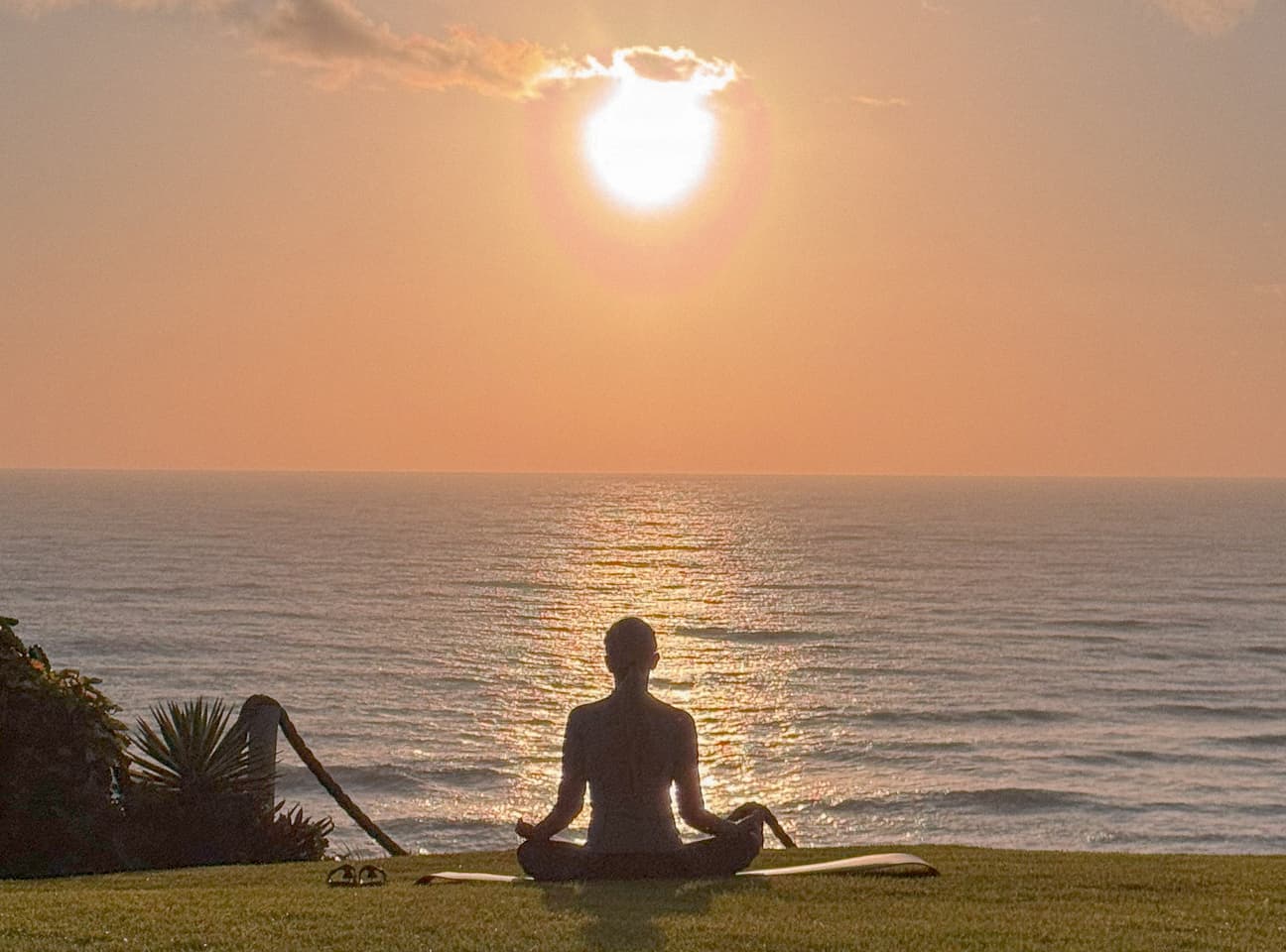 Pessoa meditando sobre um tapete de yoga no gramado diante do mar ao pôr do sol