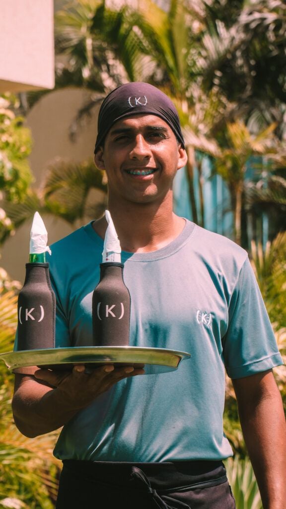Funcionário do Kilombo Villas sorrindo com bandeja de cervejas geladas em jardim tropical