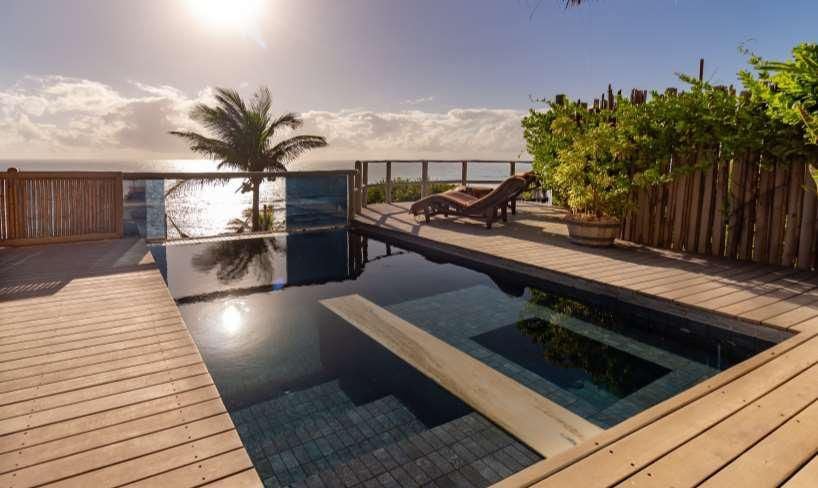 Piscina infinita escura com deck de madeira e espreguiçadeira ao pôr do sol com oceano ao fundo