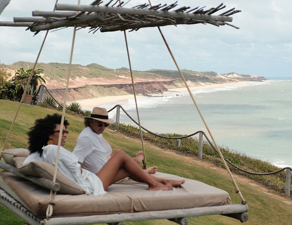 Casal descansando em uma cama suspensa de frente para o mar e as falésias da Praia da Pipa