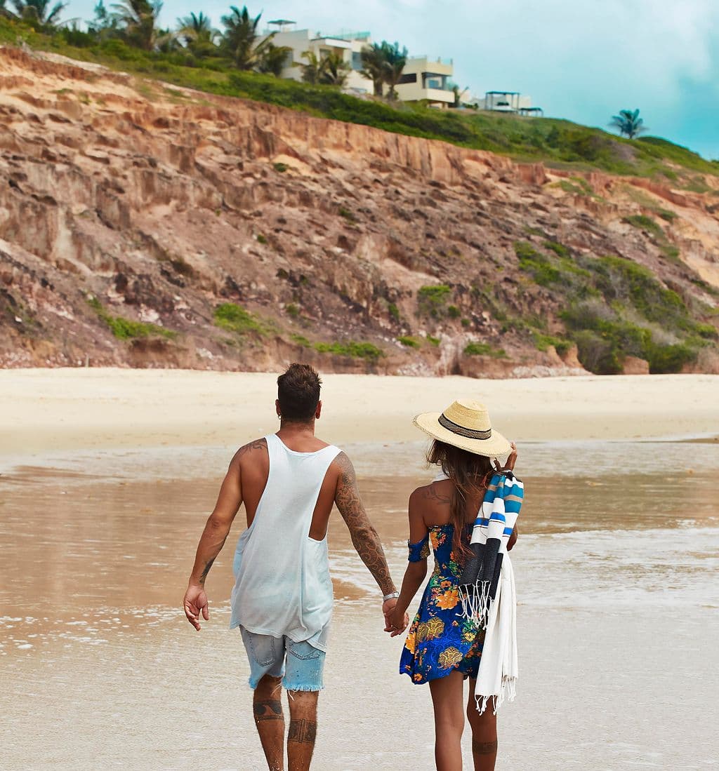 Casal caminhando de mãos dadas pela praia, com falésias ao fundo, perto de Pipa