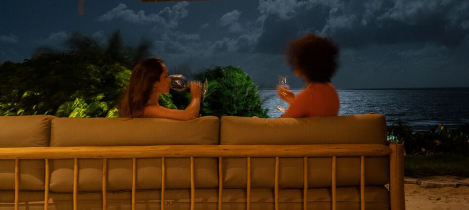 Duas amigas brindando com taças de vinho no sofá de madeira à noite com vista para o oceano
