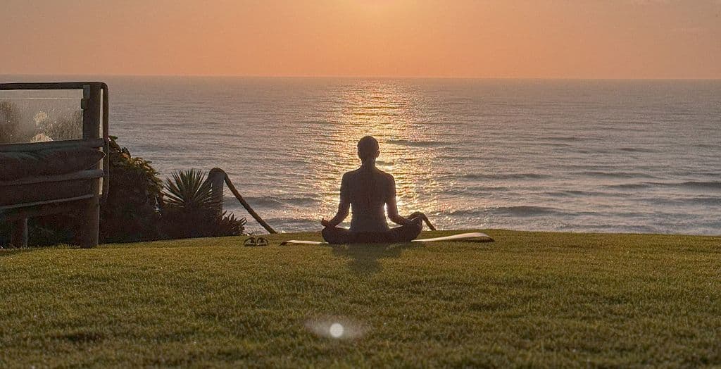 Pessoa meditando em posição de lótus no gramado do Kilombo Villas, diante do mar ao pôr do sol