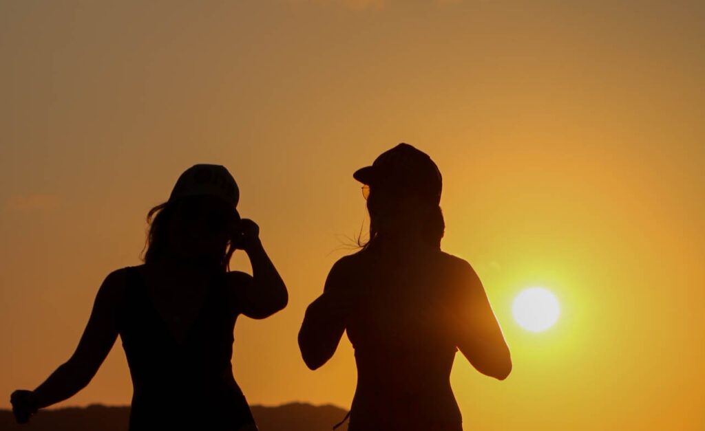 Silhuetas de duas mulheres fotografando o pôr do sol cor de laranja nas dunas