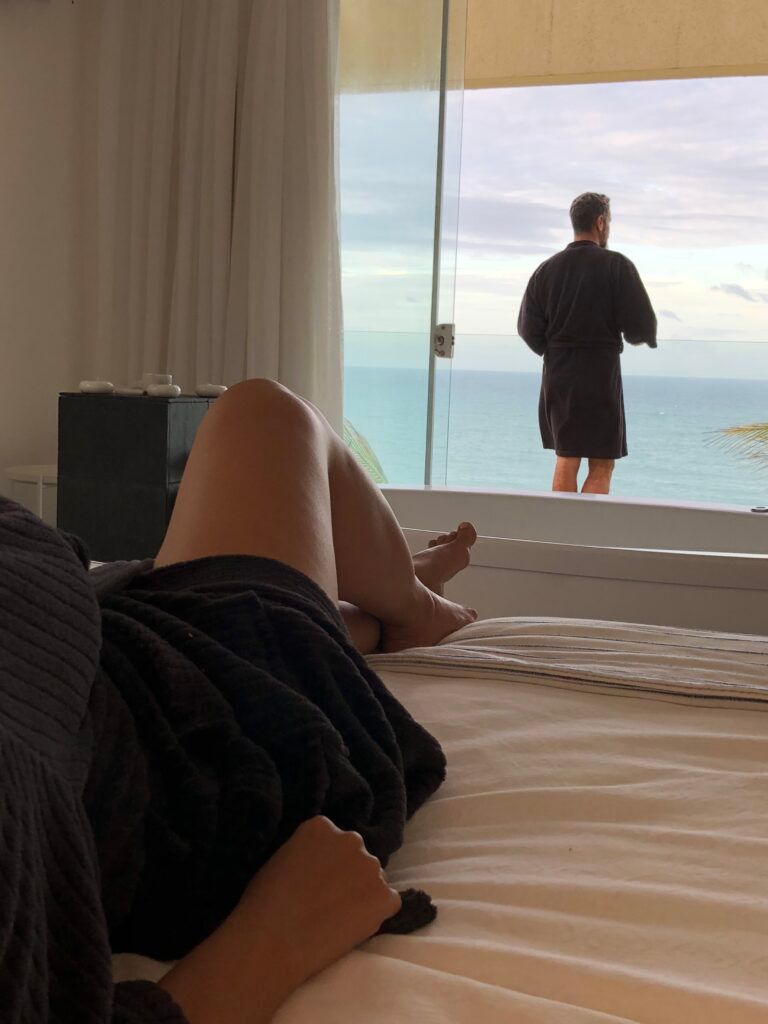 Casal de roupões escuros em suíte com homem na varanda e vista para o oceano
