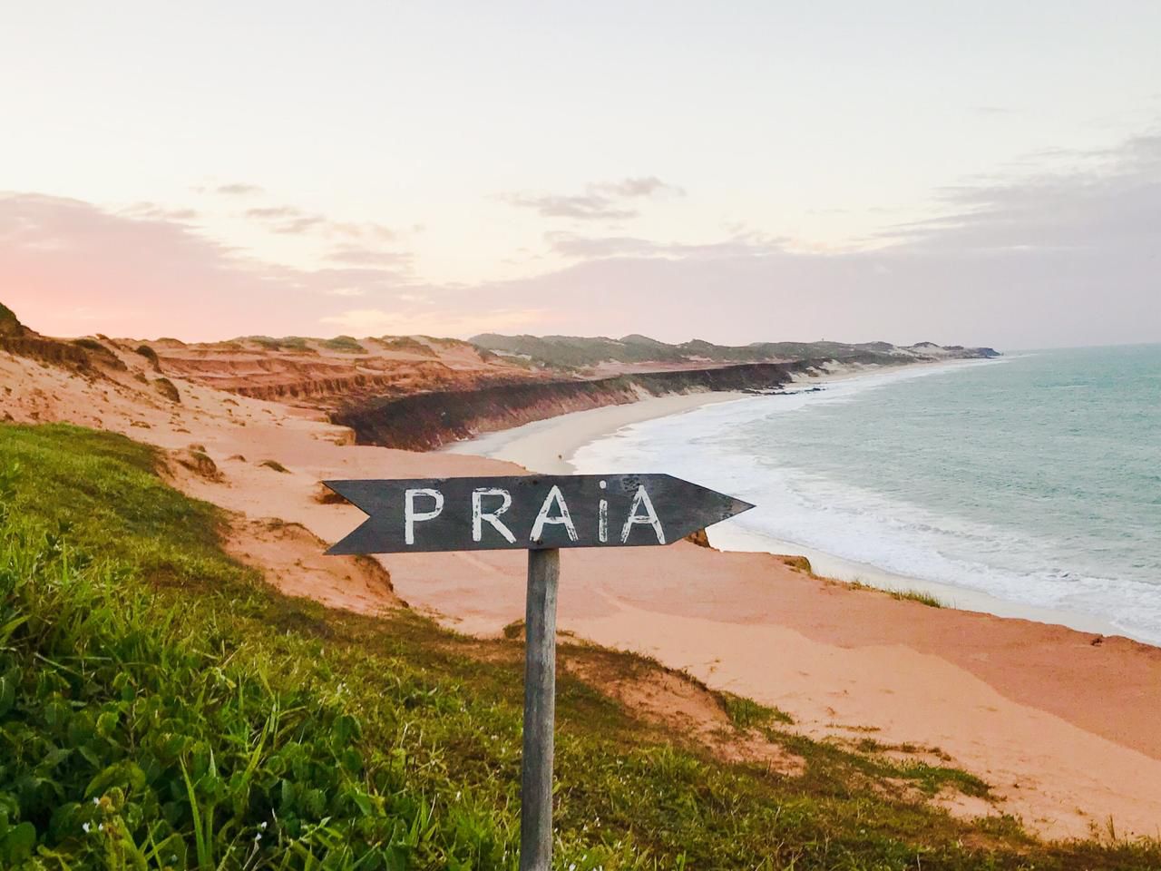 Placa de madeira "PRAIA" apontando para a praia de Sibauma com falésias avermelhadas ao fundo