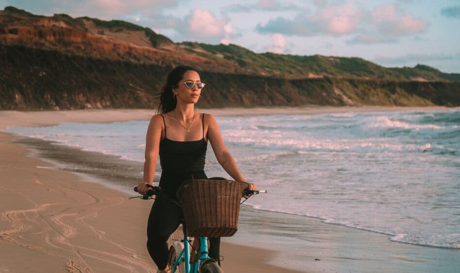 Mulher de preto pedalando bicicleta na beira-mar de Sibauma ao entardecer com falésias
