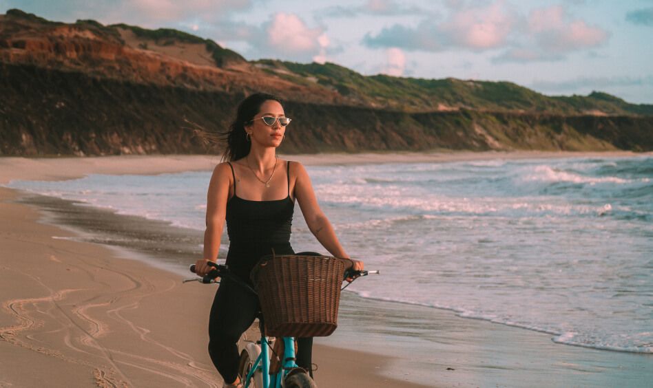 Mulher de preto pedalando bicicleta na beira-mar de Sibauma ao entardecer com falésias