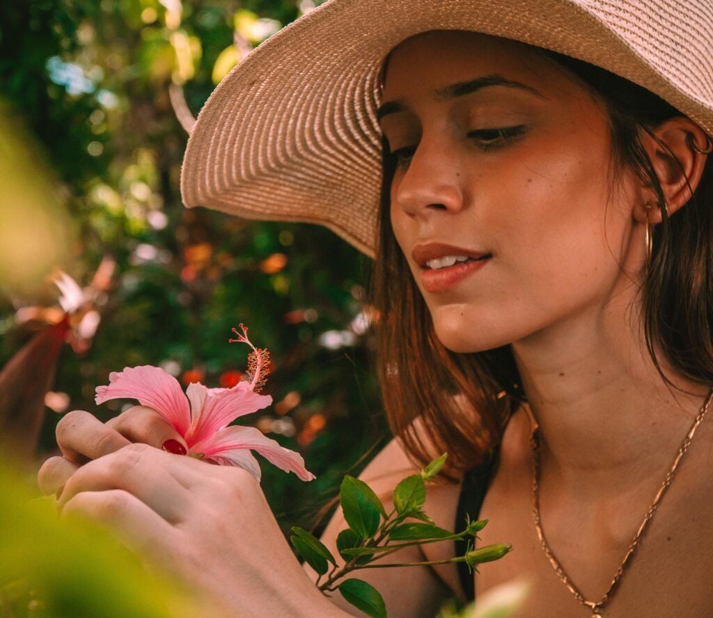 Mulher com chapéu de palha contemplando flor de hibisco rosa em jardim tropical