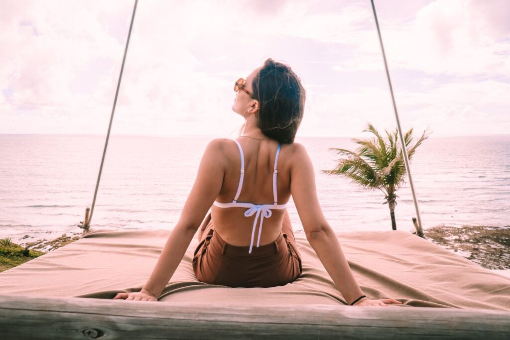 Mulher de biquíni de costas sentada em cama suspensa com óculos de sol e oceano ao fundo