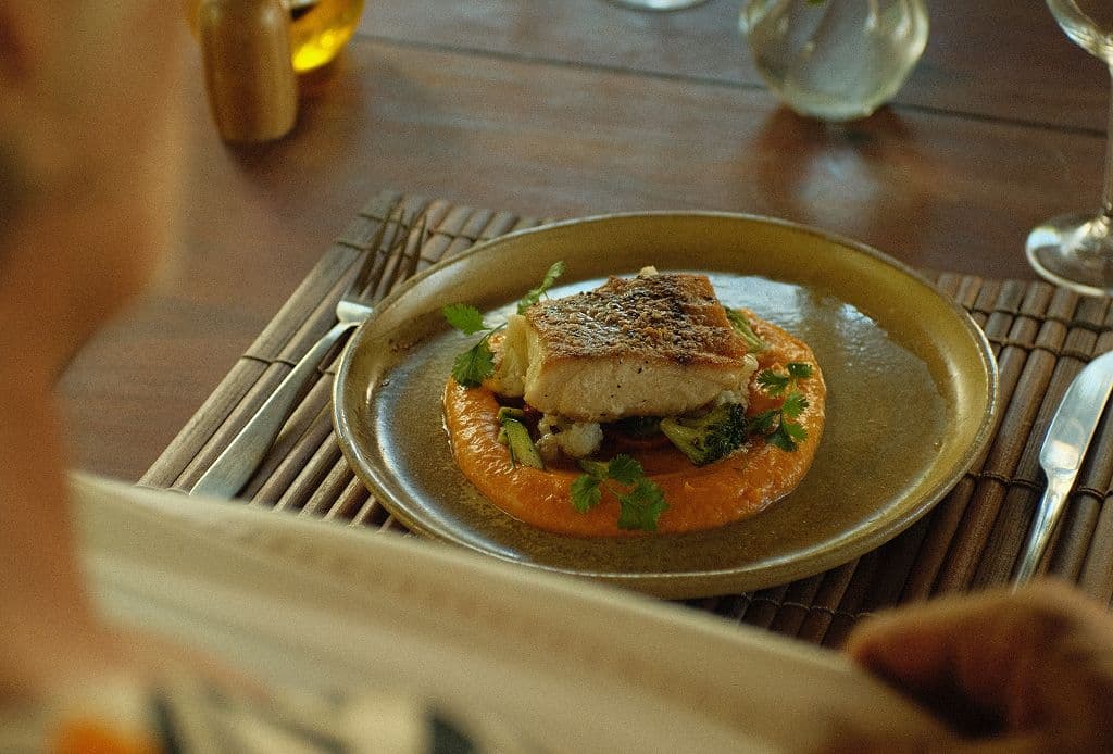Prato de peixe com purê e ervas frescas servido em mesa de madeira com vista para o mar